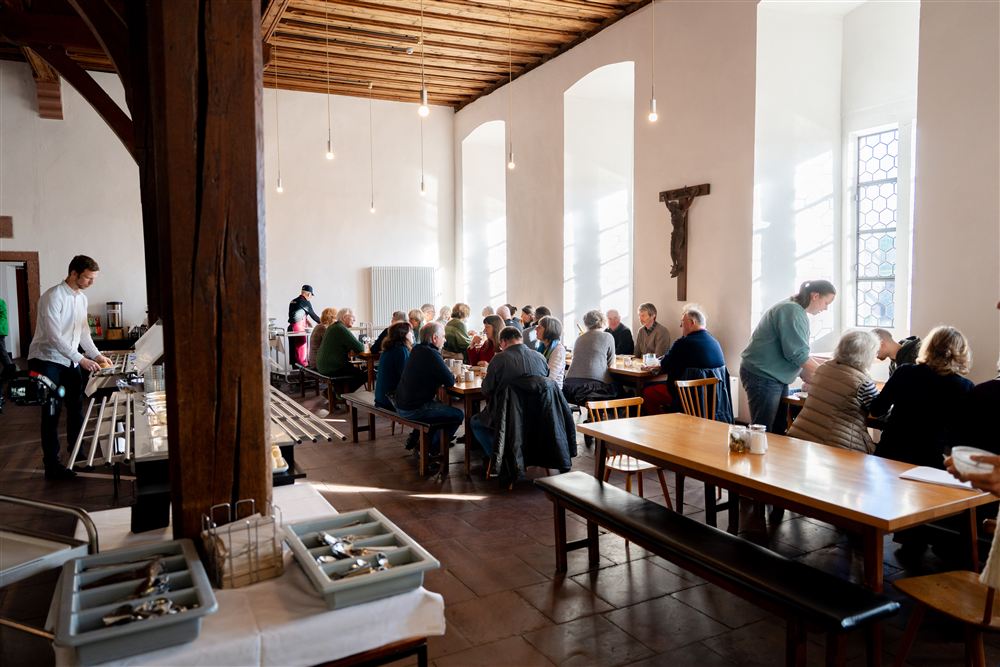 Menschen sitzen im Speisesaal beim Frühstück, die Sonne scheint stimmungsvoll durch die Fenster