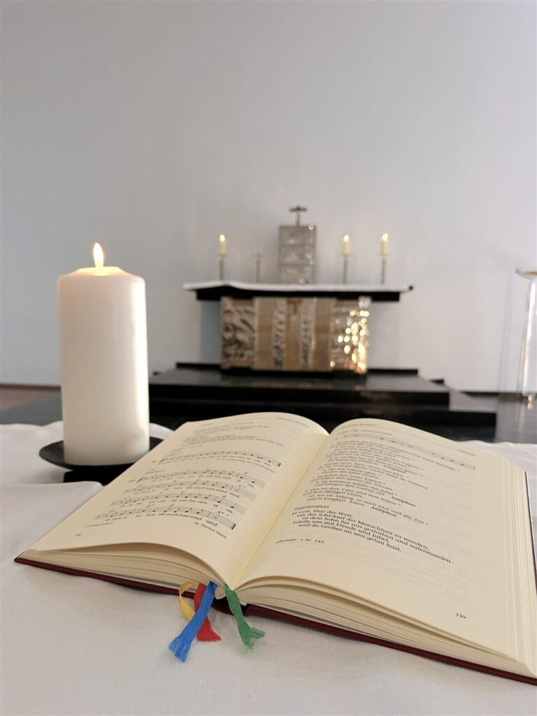 geöffnetes Liederbuch, eine Kerze daneben. Im Hintergrund ist der Altar der Burgkapelle zu sehen