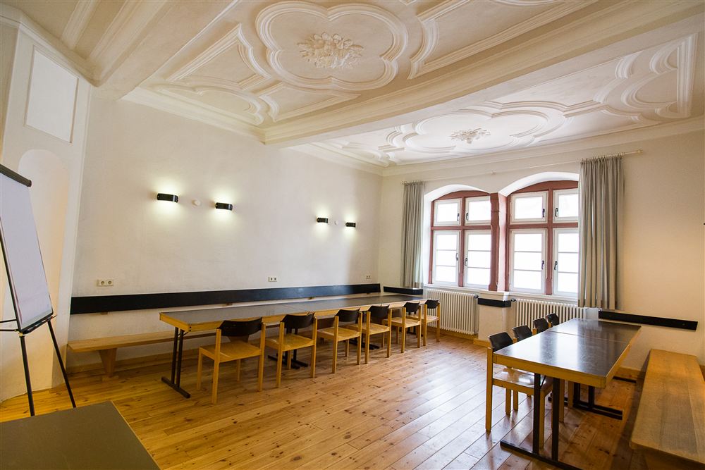 heller Tagungsraum mit barocker Stuckdecke, Holzboden, Tischen und Stühlen sowie einer Flipchart