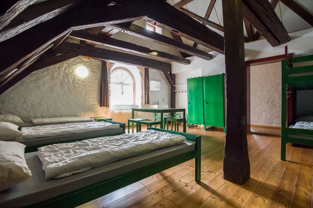 Großer Herbergs-Schlafsaal mit hoher Decke. Holzsparren sind offen sichtbar